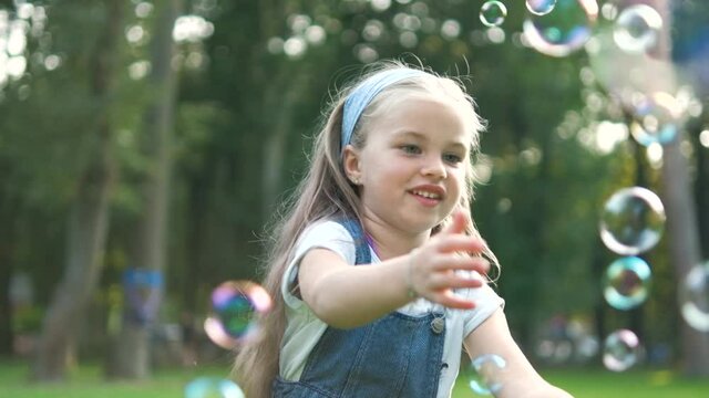 Happy little child girl catching soap bubbles outside in green park. Outdoor summer activities for children concept.