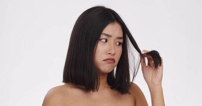 Close up portrait of upset asian woman looking at her damaged hair and pouting lips, white studio background
