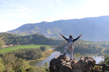 Young man standing on a rock of a cliff and enjoying the view of nature of lake and mountain in Dieng Indonesia