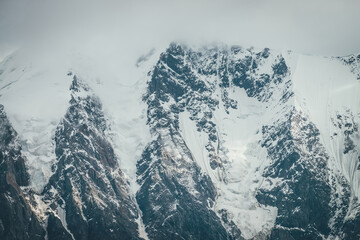 Obraz premium Atmospheric mountains landscape with big snowy mountain top with snow cornices in low clouds. Awesome minimal scenery with snow-white high pinnacle in overcast weather. Giant mountain wall in clouds.
