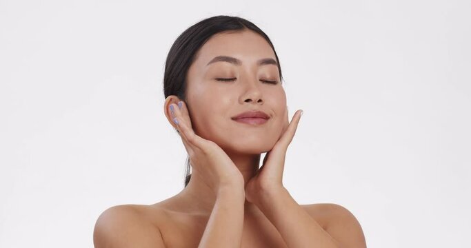 Young asian lady caressing her face skin, enjoying perfect body condition, white studio background, slow motion