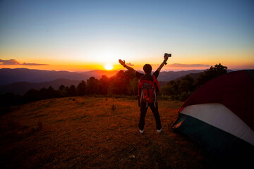 A man who loves adventure and photography. Stand on the top of the mountain with a tent, enjoy the beautiful view at sunset.