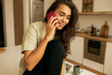Indoor image of adorable young mixed race female having conversation, drinking tea in kitchen, sitting on table with smile, listening to positive news. Cute joyful girl answering phone call