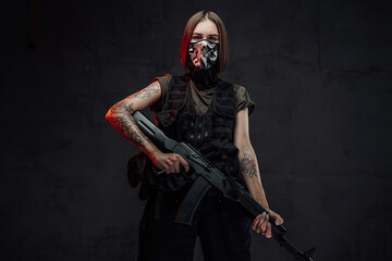 Armed with ak74 rifle and weared with mask stylish female mercenary poses in dark background.