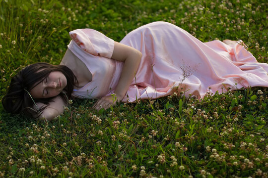 A girl with dark hair in a long pink dress lies and sleeps in the green grass with white clover flowers on a warm summer evening. Photo of beautiful sleeping woman in the fild with wildflowers.