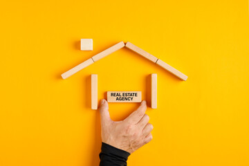 Fototapeta na wymiar Male hand placing real estate agency sign in a house made of wooden blocks on yellow background.