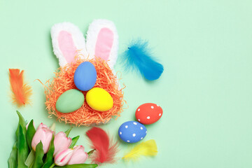 Easter eggs in a nest, bunny ears and flowers on a mint green background. Greeting card with copy space flat lay.