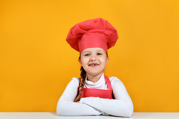 Portrait of a cute little girl chef on a yellow background. The child sits at the table and looks at the camera.