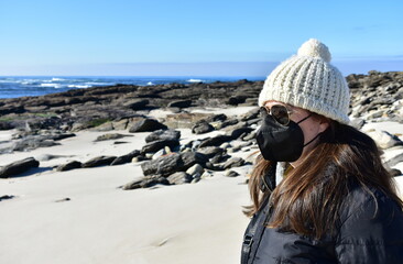 Woman with Covid-19 black face mask on a beach in Winter with black coat, sunglasses and white wool hat. KN95 or N95 or FFP2 mask.