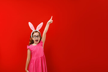 Happy cute little girl wearing easter bunny ears and glasses on red background. The child points to...