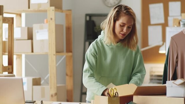 Young beautiful woman packing order into box with small gift for customer and taking notes while working in office of online clothes shop
