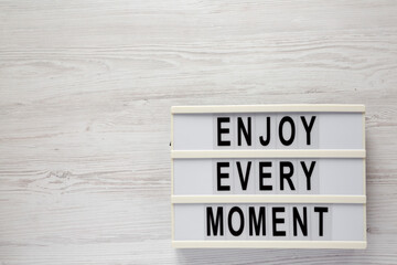 'Enjoy every moment' on a lightbox on a white wooden surface, top view. Flat lay, overhead, from above. Space for text.