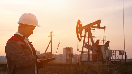 man in a white helmet with a laptop on the background of an oil rocking at sunset.