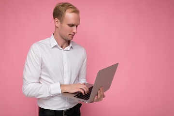 Side profile photo shot of handsome confident blonde man holding computer laptop typing on keyboard wearing white shirt looking at monitor isolated over pink background