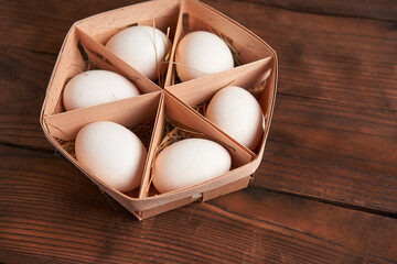White chicken eggs lie in round wooden basket which stands on a dark wooden table.