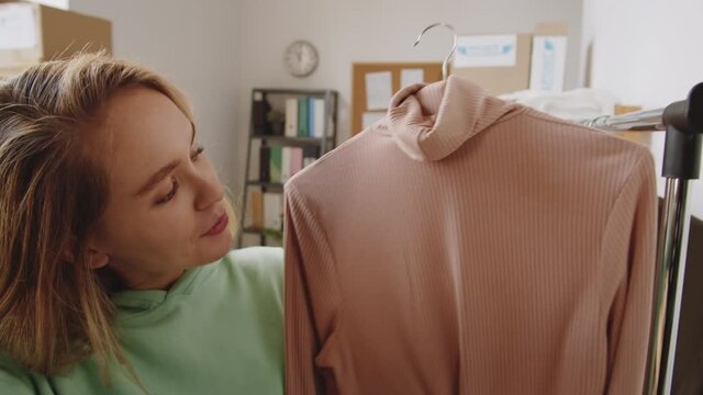 Young beautiful caucasian woman showing polo neck sweater and talking at camera via online video call while speaking with customer at work in clothes shop