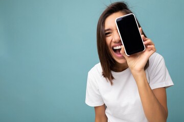 closeup Photo of pretty positive young brunette woman good looking wearing white t-shirt standing isolated on blue background with copy space holding smartphone showing phone in hand with empty screen - obrazy, fototapety, plakaty