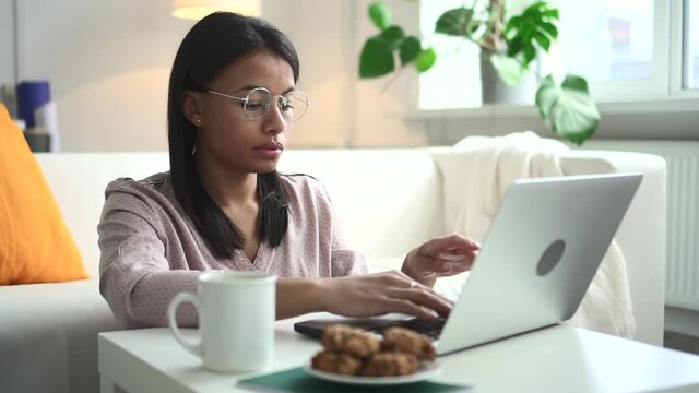 African woman with laptop at table works online at home Spbd. Female black model had glasses and beauty blouse. In background is home setting with plant and window. In room beautiful ethnic person