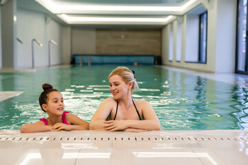 Mother and daughter in swimsuits at border of pool at the gym. They look happy, fashionable and fit.