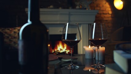 Glass of red wine, book and candle on table in dark room with fireplace. Cozy warm home in winter.