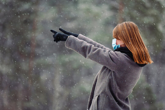 Young caucasian girl in a medical mask looks away and points with both hands with index fingers to the side against the background of the forest and small snowfalling .
