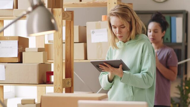 Two young multiethnic female workers using digital tablets and checking parcel boxes on shelves in online shop storage