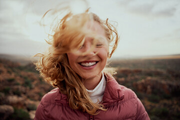 Portrait of young smiling woman face partially covered with flying hair in windy day standing at mountain - carefree woman - 406923050