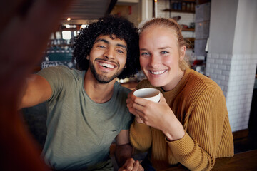 Portrait of smiling man with girlfriend holding coffee cup taking selfie in modern cafe