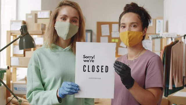 Portrait of two multiethnic female coworkers in protective gloves and masks holding poster with Sorry! WeвЂ&trade;re closed sign and posing for camera while closing store due to covid-19 lockdown