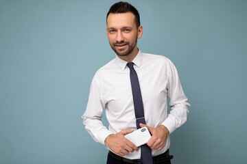 Photo of handsome good looking brunet unshaven man with beard wearing casual white shirt and tie isolated on blue background with empty space holding in hand mobile phone looking at camera