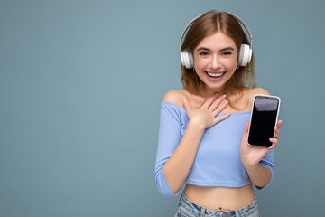 Attractive positive smiling young woman wearing stylish casual outfit isolated on colourful background wall holding and showing mobile phone with empty screen for cutout wearing white bluetooth