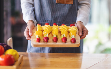 Closeup image of a female chef holding and showing a wooden plate of fresh mixed fruits on skewers...