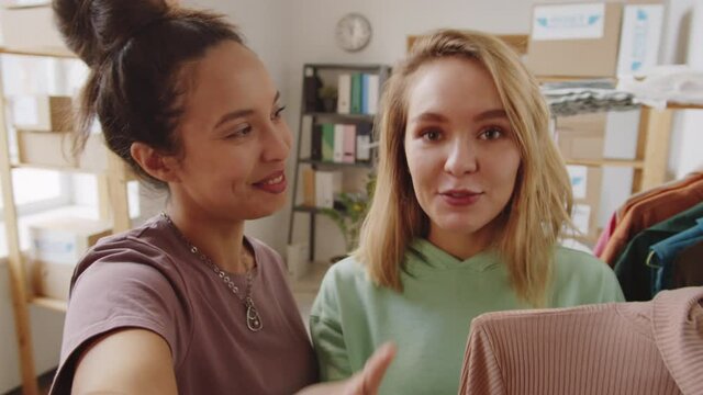 Two young multiethnic female colleagues smiling, waving and telling about polo neck sweater at camera via online video call while working remotely in clothes shop