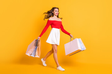 Full length body size photo girl smiling looking blank space stepping with packages from store isolated on bright yellow color background