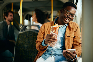 Happy black man eating a sandwich and using smart phone while commuting by bus.