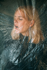 Fototapeta na wymiar Female abuse. Defocused art portrait. Domestic violence. Disturbed hurt blonde woman face with closed eyes isolated behind wrinkled texture transparent polyethylene film on dark background out of