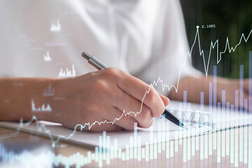 A woman trader in formal wear writing down some ideas to research stock market to proceed right investment solutions. Wealth management concept. Hologram Forex chart over close up shot.