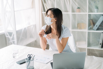 Pandemic furlough. Office relocation. Unemployment crisis. Pensive sad female employee in protective face mask thinking on new project at light workplace with laptop covered furniture.
