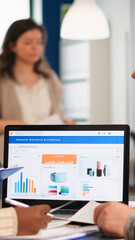 Busy multicultural, diverse employees analysing annual financial statistics sitting at desk in front of laptop holding documents searching business solutions. Team of businesspeople working in company