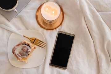 Smartphone with empty screen on the bed with cinnamon bun and burning candle