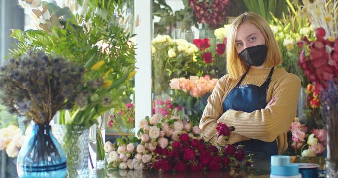 Medium shot of woman in medical mask working in flower store. Female florist standing in blossom bouquet background, crossing arms and looking in camera. Designing, floral workshop, working concept.