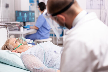 Obraz premium Doctor checking senior woman with oxygen mask breathing hard because of infection with covid19. Patient in hospital room laying on medical bed with infected lung during coronavirus pandemic having