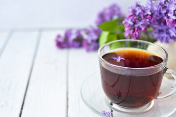 Delicate setting of the morning tea table with lilac flowers, a transparent cup of tea and a saucer and a white vase on a white wooden board. Copy space. Spring breakfast concept