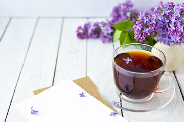 Delicate morning tea table setting with lilac flowers, a transparent cup of tea and a saucer and a white vase, white and craft note paper on a white wooden board. Mockup. 