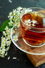 A transparent cup of teapot tea with a saucer and a white bird cherry flower on a black background. Copy space. Spring breakfast