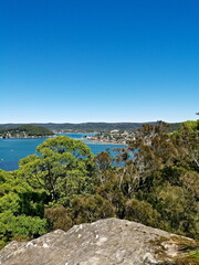 Fototapeta na wymiar Beautiful view of a bay full of boats with tall tree in the foreground and mountains, trees and deep blue sky in the background, Allen Strom Lookout, Brisbane Water, Rocky Point Trail, New South Wales