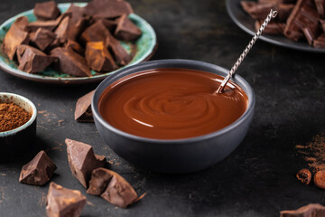 Melted organic chocolate in a bowl with a spoon and pieces of dark chocolate on a dark textured background. Confectionery home concept.