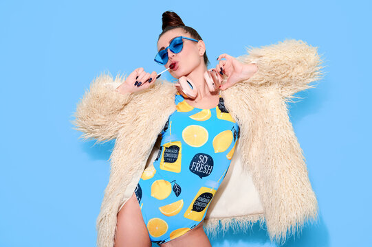 Girl dj wearing swimsuit and fur coat lick lollipop on bright blue background