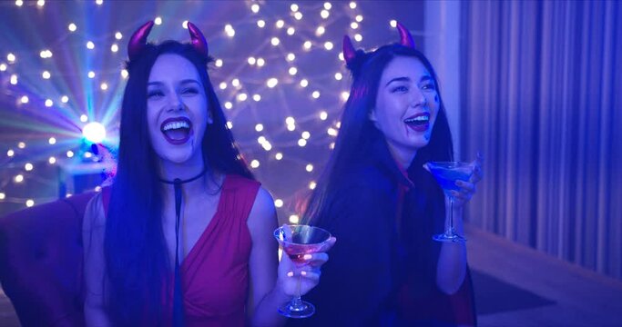 Medium Shot, Handheld - Two beautiful seductive young Caucasian female friends, dressing in she devils drinking and dancing at a Halloween costume party in a dark room with decorative lights.