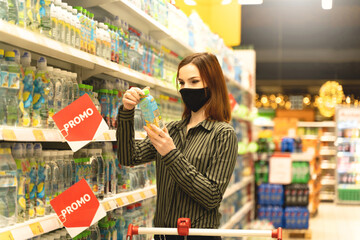 Woman with mask in supermarket makes grocery purchases. Customer read composition of product in hypermarket. Girl chooses drink. Shopper in store